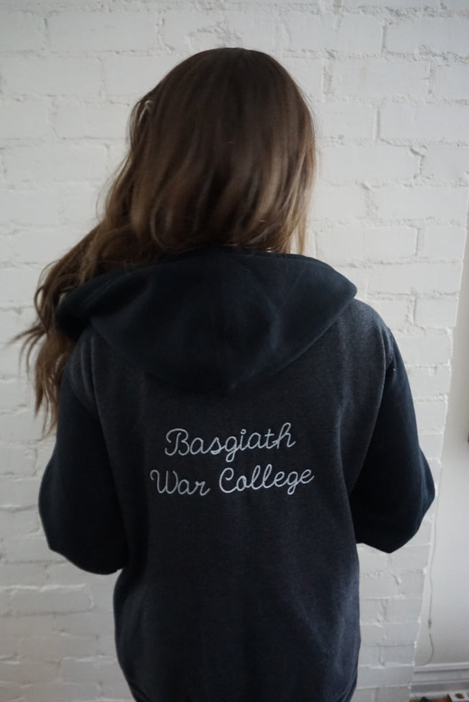 Person wearing a dark hoodie with 'Basgiath War College' text against a white brick wall.